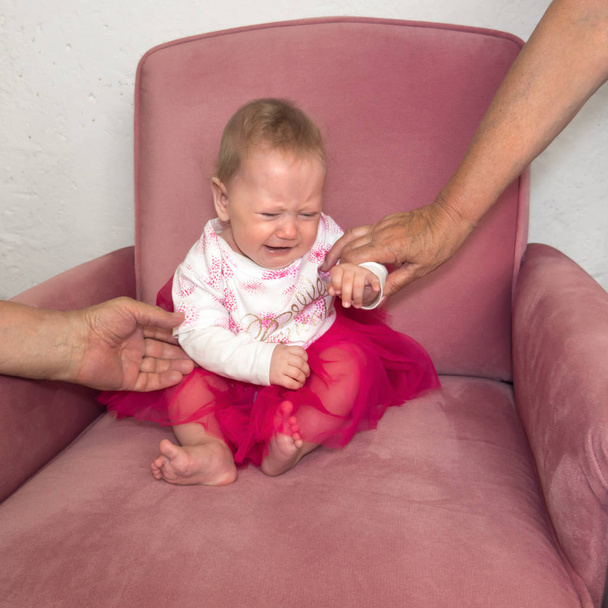 Bebê chorando sentado na cadeira. Mãos de avó segurando criança sentada na cadeira. Emoção infantil negativa. Expressão facial. Retrato de menina vestida sentada na cadeira
. - Foto, Imagem