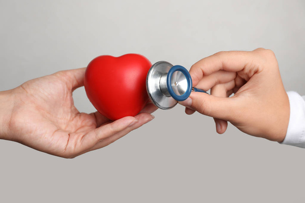 Врач держит стетоскоп возле игрушечного сердца в руке пациента на сером фоне, крупным планом. Проверка пульса
 - Фото, изображение
