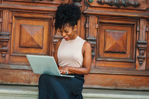 Δρόμο προς την επιτυχία. Νέοι αφρικανική αμερικανική γυναίκα με Άφρο hairstyle φορώντας κορυφή αμάνικο ελαφρύ χρώμα, μαύρο σκετς, κάθονται από πόρτα vintage γραφείου στη Νέα Υόρκη, που εργάζεται στο φορητό υπολογιστή, χαμογελώντας - Φωτογραφία, εικόνα
