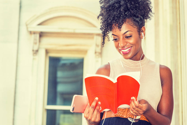 Молодая счастливая афроамериканка, студентка колледжа с афро-прической сидит у винтажного офисного здания в Нью-Йорке, читает красную книгу, слушает музыку с наушником и сотовым телефоном, улыбается
 - Фото, изображение