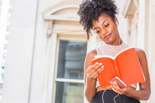 Young gelukkig African American vrouwelijke student met afro kapsel zitten door de vintage stijl kantoorgebouw in New York, lezen van rode Boek, luisteren van muziek met oortelefoon en mobiele telefoon, glimlachend - Foto, afbeelding