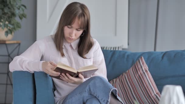 Dívka čtení kniha zatímco sedí na gauči - Záběry, video