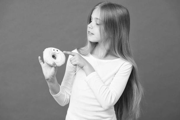 Girl holding fluffy doughnut. Glazed bagel with eyes, creative dessert. Kid eating icing out off doughnut, sweet tooth heaven. Child with long hair wearing white jumper isolated on purple background - Foto, imagen