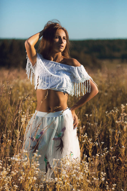 Attractive young woman with slim athletic body standing posing in field of tall grass on sunset with hand in her hair - Valokuva, kuva