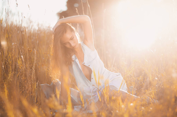 Attractive young woman in jean sundress sitting in a field of tall grass and reading a book under summer sunlight beam - Foto, afbeelding