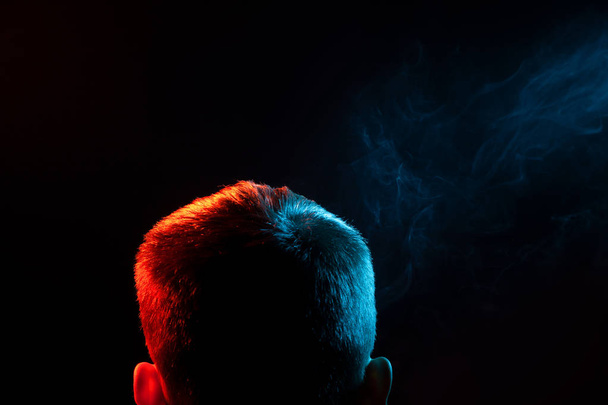 Blick von hinten auf den Kopf eines Mannes im Hemd, der einen Dampf raucht und auf einem schwarzen, isolierten Hintergrund mehrfarbigen grünen Rauch aus verschiedenen Richtungen von sich selbst ausatmet. die Gedanken und die Seele des Menschen. - Foto, Bild