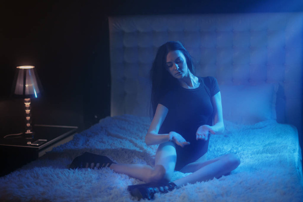 sexy junge Frau mit schwarzen Haaren und nackten langen Beinen sitzt auf weißem Bett und spielt mit einem magischen Mondlicht, das auf ihre Hände fällt, und trägt sexy schwarze Dessous als Hexenkostüm auf einer hilflosen Party - Foto, Bild