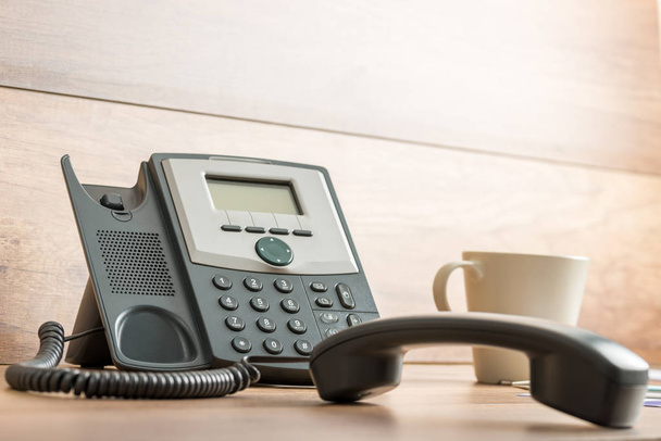 Teléfono fijo negro con teléfono desconectado en escritorio de madera y taza de café al lado
. - Foto, Imagen