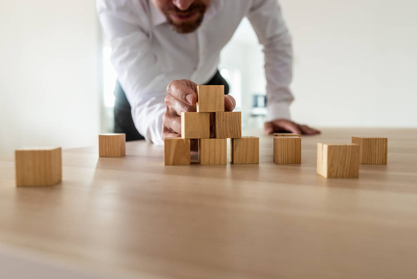 Businessman leaning in to carefully assemble pyramid shape with blank wooden blocks on office desk. Conceptual of business start up and  vision. - Photo, Image