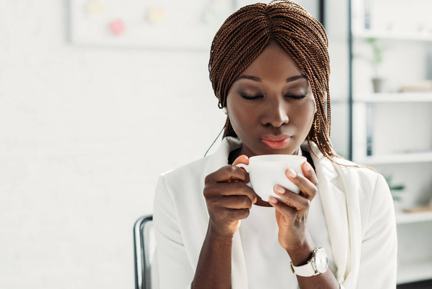 beautiful african american adult businesswoman in white suit with eyes closed sitting at desk, holding cup of coffee and inhaling aroma - Fotografie, Obrázek