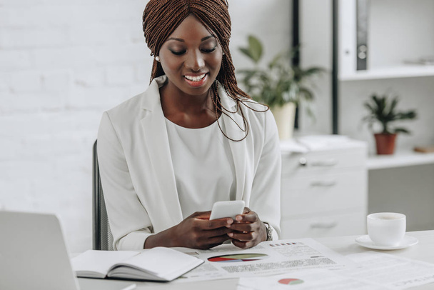 улыбающаяся африканская взрослая американская бизнесвумен сидит за столом, работает и смотрит на смартфон
 - Фото, изображение