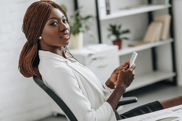beautiful african american adult businesswoman in white formal wear sitting at desk, looking at camera and using smartphone in office - Fotografie, Obrázek