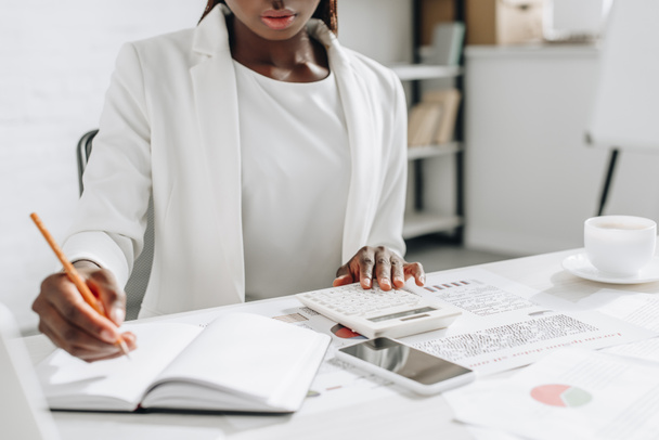 vista recortada de la mujer de negocios adulta afroamericana en ropa formal blanca escribiendo en cuaderno y trabajando en escritorio de oficina
 - Foto, Imagen