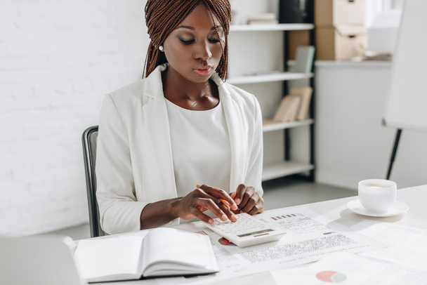 白のフォーマルな服装のオフィスの机で働いて、電卓を使用しての焦点をアフリカ系アメリカ人大人女性実業家 - 写真・画像
