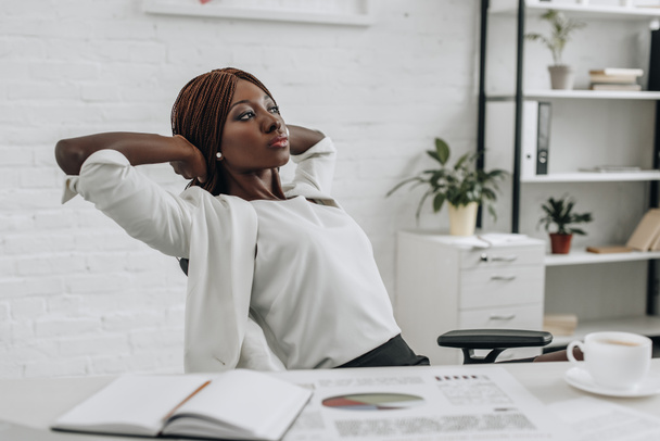 mujer de negocios adulta afroamericana en ropa formal blanca con las manos detrás de la cabeza sentadas en el escritorio y relajándose en la oficina moderna
 - Foto, imagen