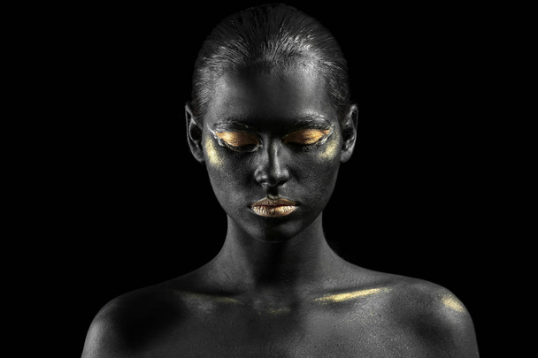 Mooie vrouw met zwarte en gouden verf op haar lichaam tegen donkere achtergrond - Foto, afbeelding