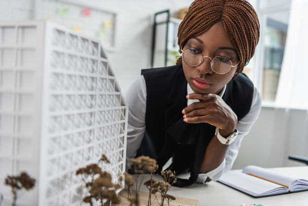 серьезная африканская женщина-архитектор в очках и формальной одежде трогательный подбородок рукой и работает над строительным проектом
 - Фото, изображение