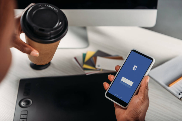 vista recortada de mujer de negocios afroamericana sosteniendo café para llevar y teléfono inteligente con aplicación de Facebook en la pantalla
 - Foto, imagen