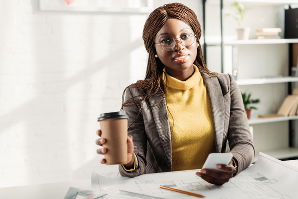 Afryki amerykański architekt kobiece w okularach trzyma kawę iść, za pomocą smartfona i pracę przy biurku na projekt z odbitek - Zdjęcie, obraz