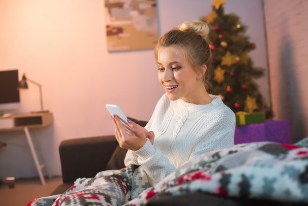 happy young blonde woman sitting on couch and using smartphone at christmas time - Photo, Image