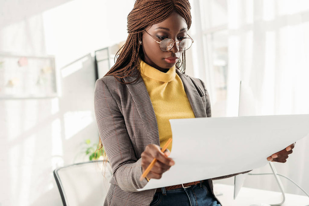 focalizzata afro-americana architetto donna in occhiali che tengono cianografia e matita in ufficio
 - Foto, immagini