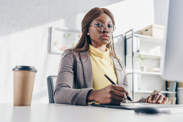 konzentrierte afrikanisch-amerikanische Geschäftsfrau sitzt am Computer-Schreibtisch, bedient sich eines Grafik-Tablets und blickt am Arbeitsplatz in die Kamera - Foto, Bild