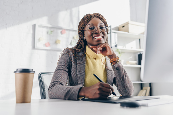 sonriente afroamericana adulto mujer de negocios mirando a la cámara, sentado en el escritorio de la computadora y el uso de la tableta gráfica
 - Foto, imagen