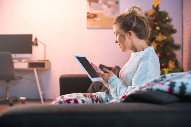side view of young blonde woman sitting on couch and using tablet at christmas time  - Foto, Imagem