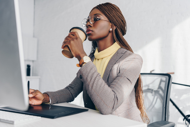 beautiful confident african american businesswoman using graphics tablet and drinking coffee to go at workplace - Photo, Image