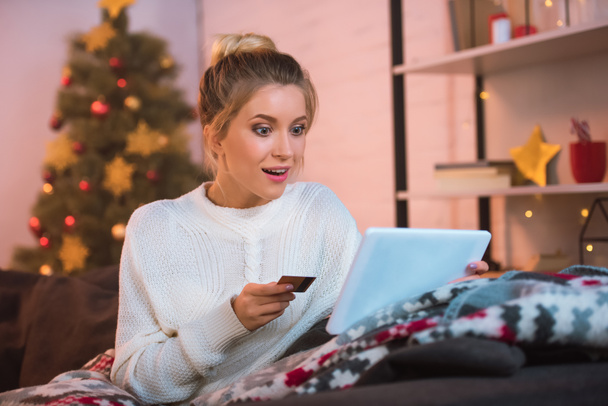 surprised young blonde woman sitting on couch holding credit card and using digital tablet for online shopping at christmas time - Photo, Image