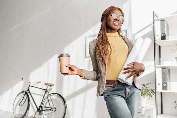smiling african american adult female architect in glasses holding coffee to go and blueprints at workplace - Фото, изображение