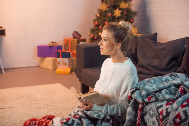 Chica encantadora sonriendo y leyendo libro en casa en la víspera de Navidad
 - Foto, Imagen