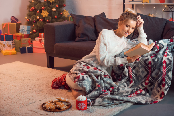 Очаровательная девушка улыбается и читает книгу дома в канун Рождества
 - Фото, изображение