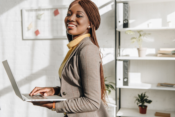 Afrikanisch-amerikanische erwachsene Geschäftsfrau hält Laptop am Arbeitsplatz, lächelt und blickt in die Kamera - Foto, Bild