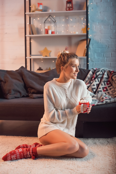 atractiva mujer sentada en calcetines con dibujos navideños en la alfombra con taza de cacao
 - Foto, Imagen
