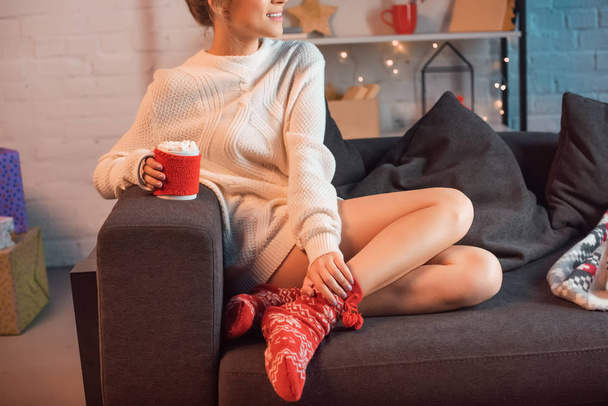 partial view of smiling young blonde woman sitting on couch and holding cup of hot cocoa with marshmallows at christmas time  - Fotó, kép