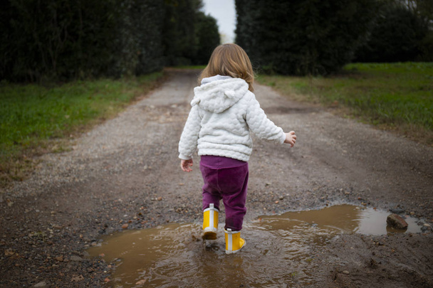 Kleinkind, Rückansicht, plätschernd in Pfützen auf schlammiger Landstraße mit gelben Regenstiefeln. Thema im Mittelpunkt. - Foto, Bild