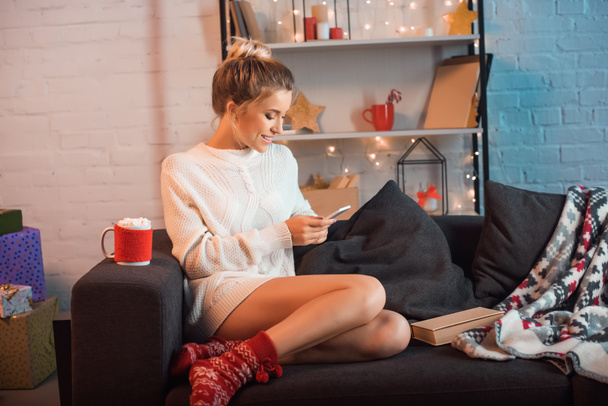 smiling young blonde woman sitting on couch and using smartphone at christmas time  - Fotó, kép