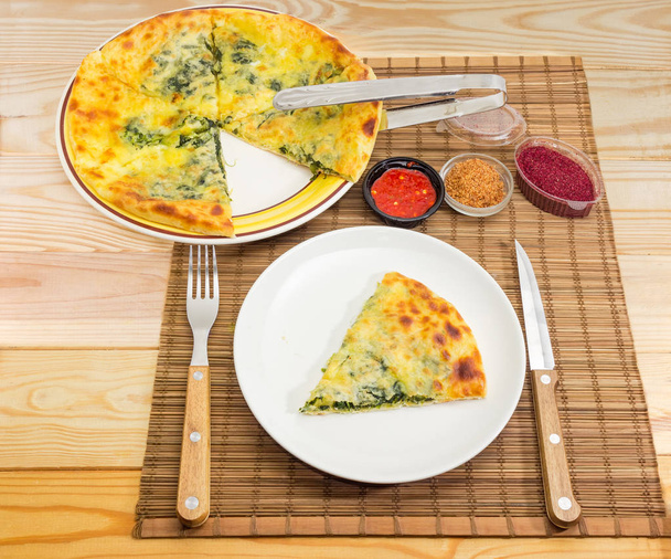 Partly sliced khachapuri on dish, one slice on other dish, fork, knife and seasonings on a bamboo place mat on a wooden light colored rustic table - Foto, Imagem