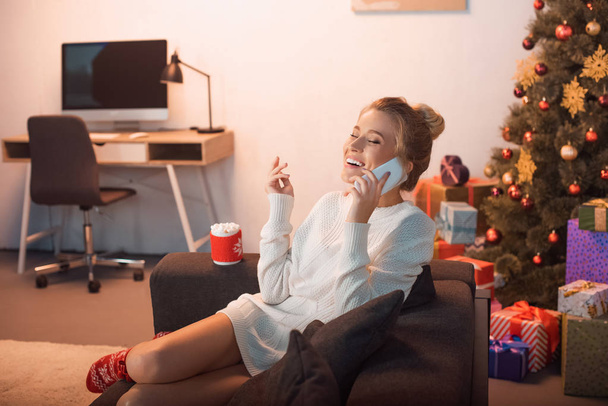ridendo giovane donna bionda seduta sul divano e parlando sullo smartphone nel periodo natalizio
  - Foto, immagini