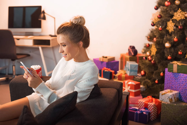 smiling young blonde woman sitting on couch and using smartphone at christmas time  - Photo, Image