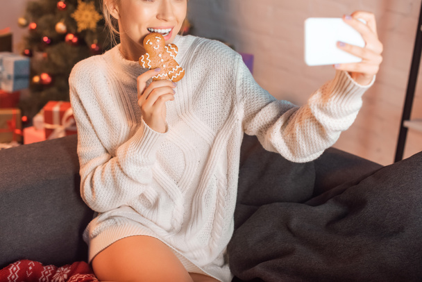 young blonde woman posing with gingerbread cookie and taking selfie on smartphone at christmas time - Photo, Image