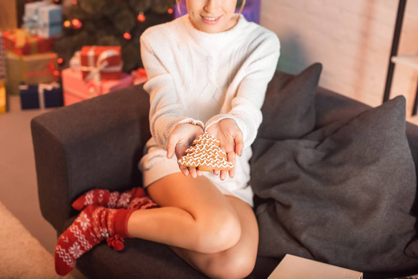 cropped view of smiling young woman sitting on couch and holding gingerbread cookie in outstreched hands at christmas time - Photo, Image