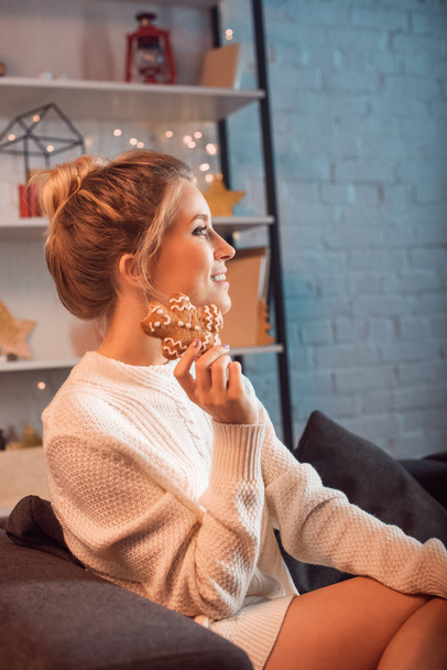 smiling young blonde woman sitting on couch, posing and holding gingerbread cookie at christmas time - Zdjęcie, obraz