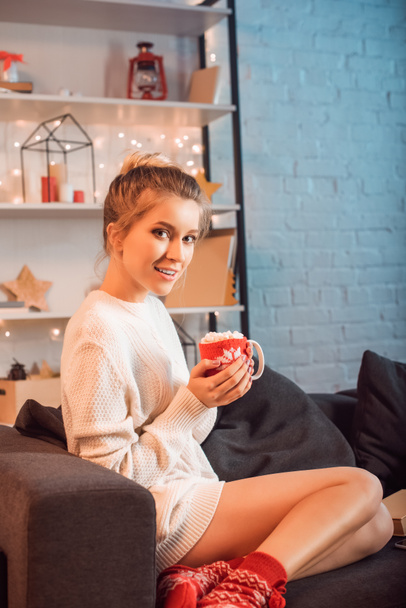 attractive young blonde woman sitting on couch and holding cup of hot cocoa with marshmallows at christmas time - Photo, Image