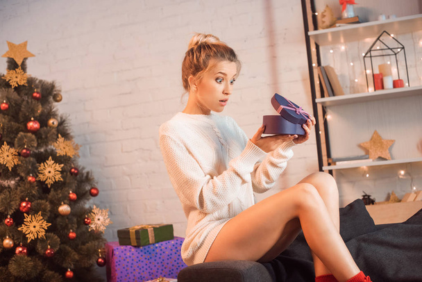 surprised young blonde woman sitting on couch and opening present at christmas time  - Фото, изображение