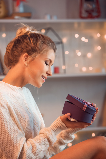 Lächelnde junge blonde Frau öffnet Geschenk zur Weihnachtszeit mit verschwommenem Hintergrund - Foto, Bild