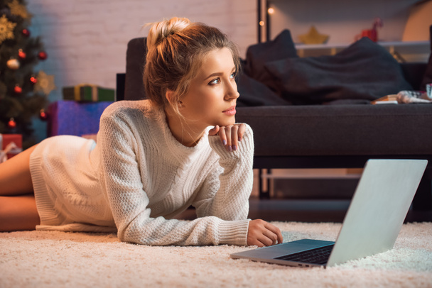 Nachdenkliche junge blonde Frau liegt zur Weihnachtszeit mit Laptop am Boden  - Foto, Bild