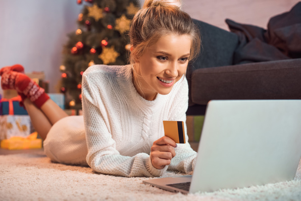 smiling young blond woman lying on floor, holding credit card, using laptop and doing online shopping at christmas time  - Photo, Image