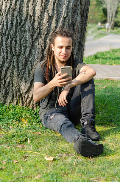 Młody człowiek patrzy na jego telefon komórkowy pod drzewem. - Zdjęcie, obraz